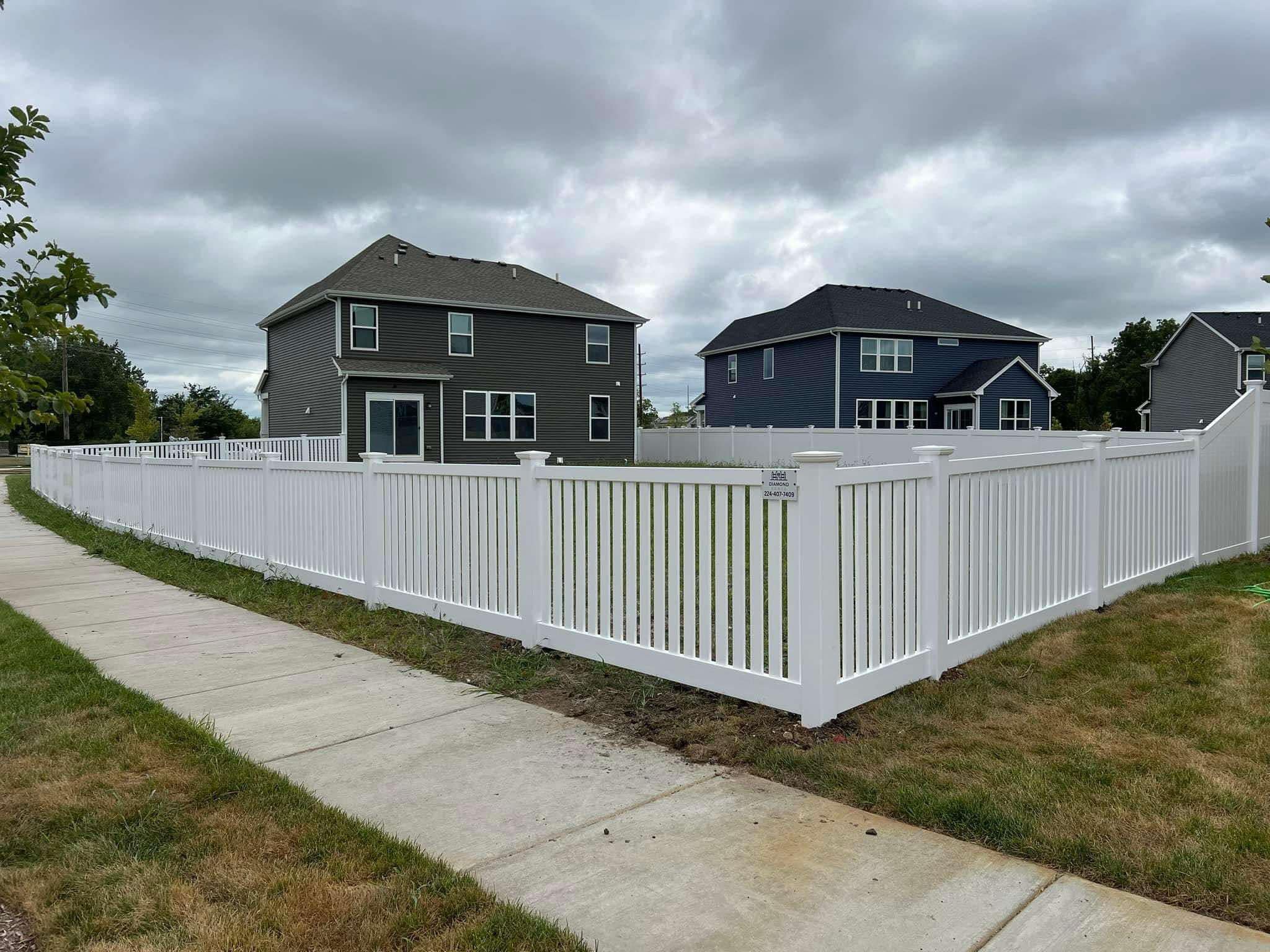 Modern white vinyl picket-style fence wrapping around new suburban homes under cloudy sky — combines elegance and durability; features Diamond Fence sign (224-407-7409) along sidewalk