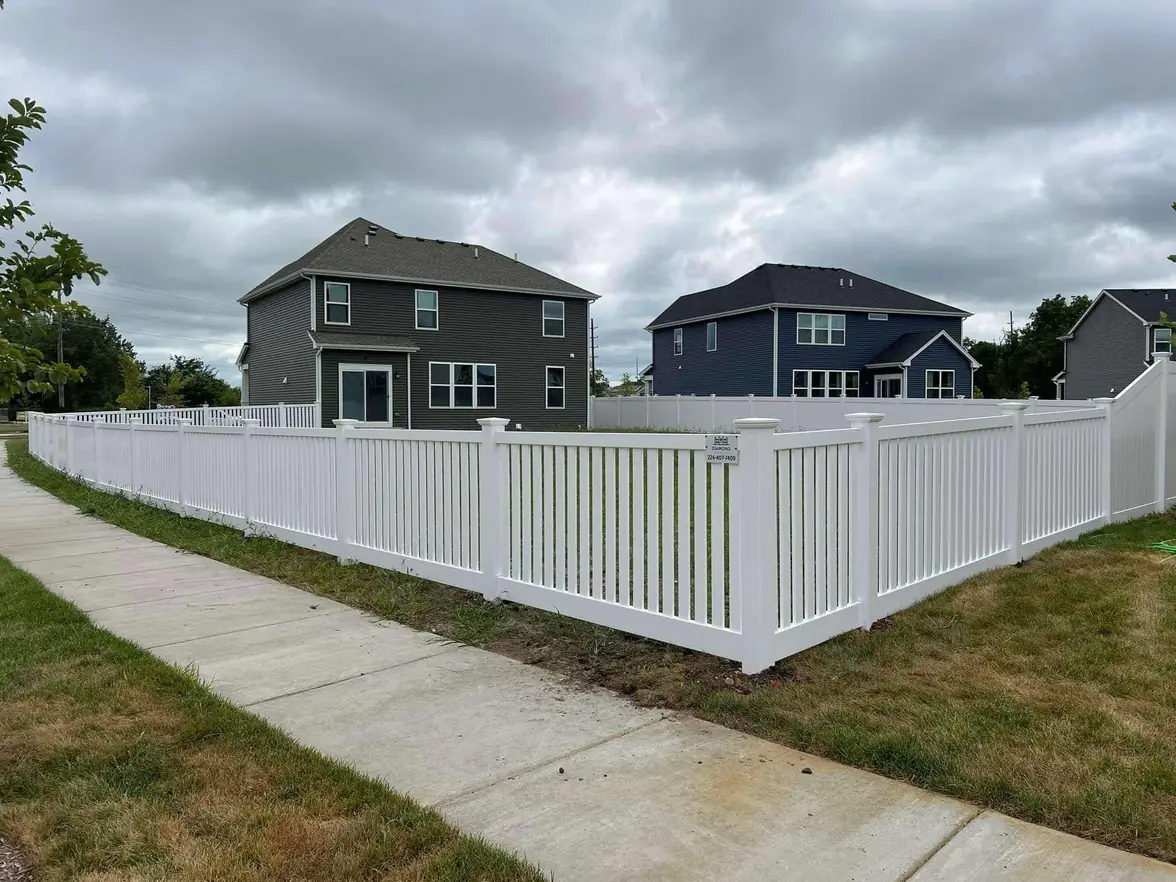 Semi-open white vinyl fence around a corner lot beside a sidewalk, with two-story houses behind
