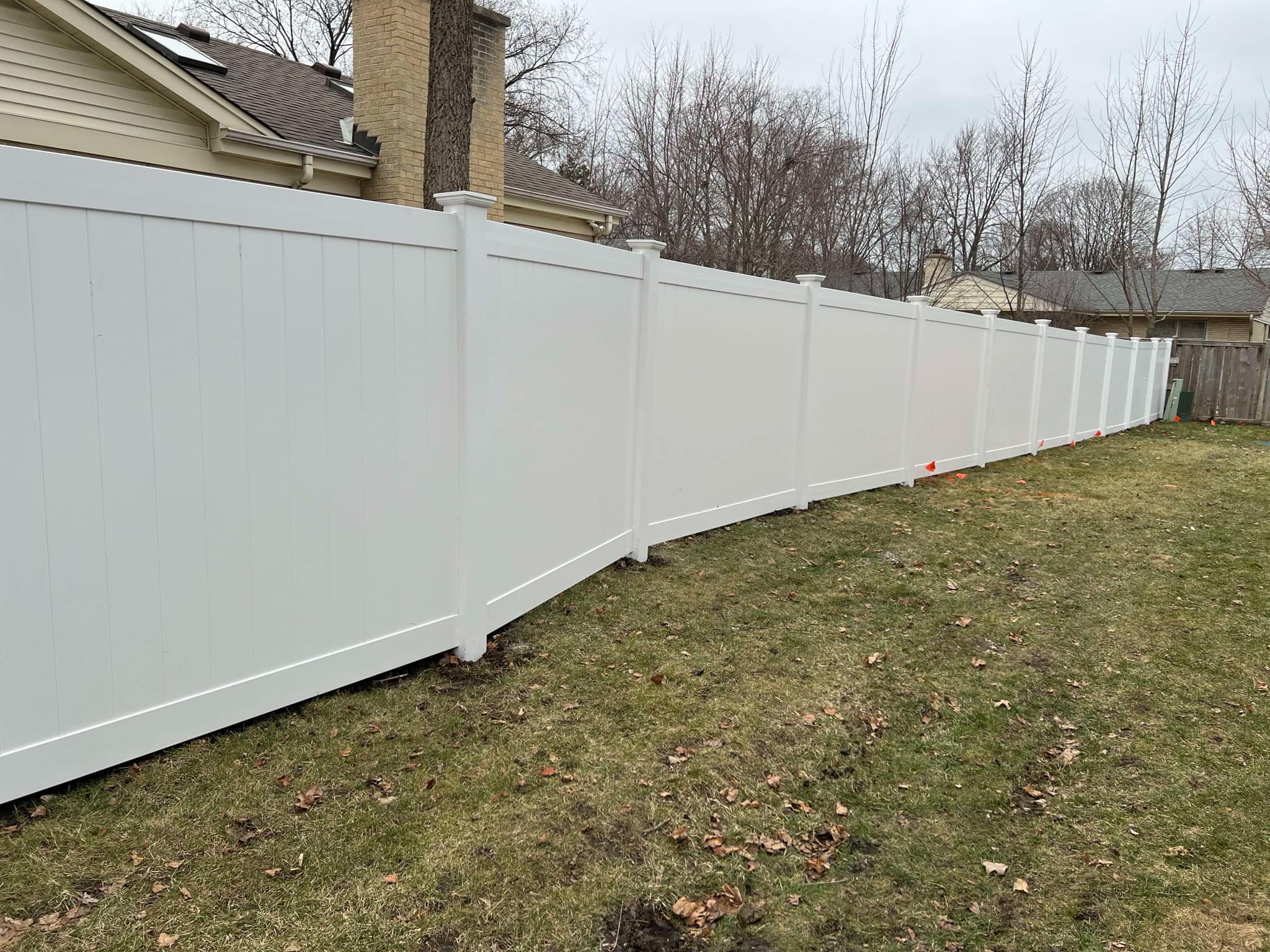 Long run of newly installed white vinyl privacy fence following property line beside beige home with brick chimney — clean lines, no gaps, and professional finish on dormant lawn